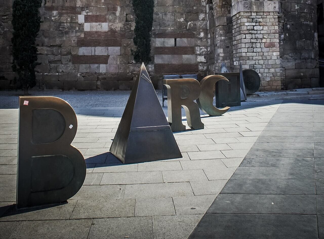 Escultura urbana con la palabra “Barcino” formada por letras grandes de color rojo, instalada en una plaza de Barcelona, obra de Joan Brossa, con edificios históricos al fondo; fotografía de Los Paseos.