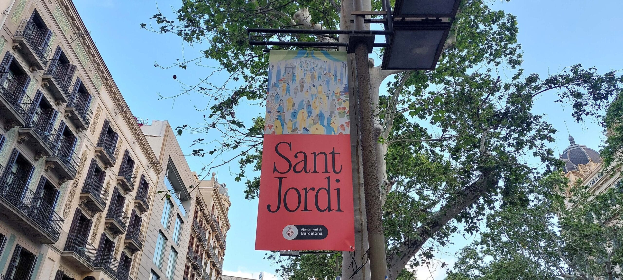 Cartel anunciador de la festividad de Sant Jordi en La Rambla de Barcelona, integrado en el espacio urbano durante la celebración. La imagen muestra la presencia de la tradición literaria y floral en una de las principales avenidas de la ciudad.